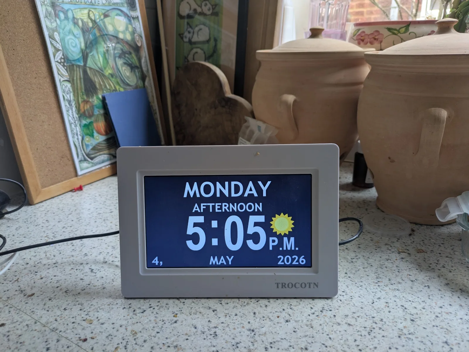 A TROCOTN dementia clock on a kitchen worktop, displaying 'MONDAY AFTERNOON 5:05 P.M.' with a sun icon and the date '4, MAY 2026' in large white type on a blue background, with terracotta storage jars and a framed picture behind it.