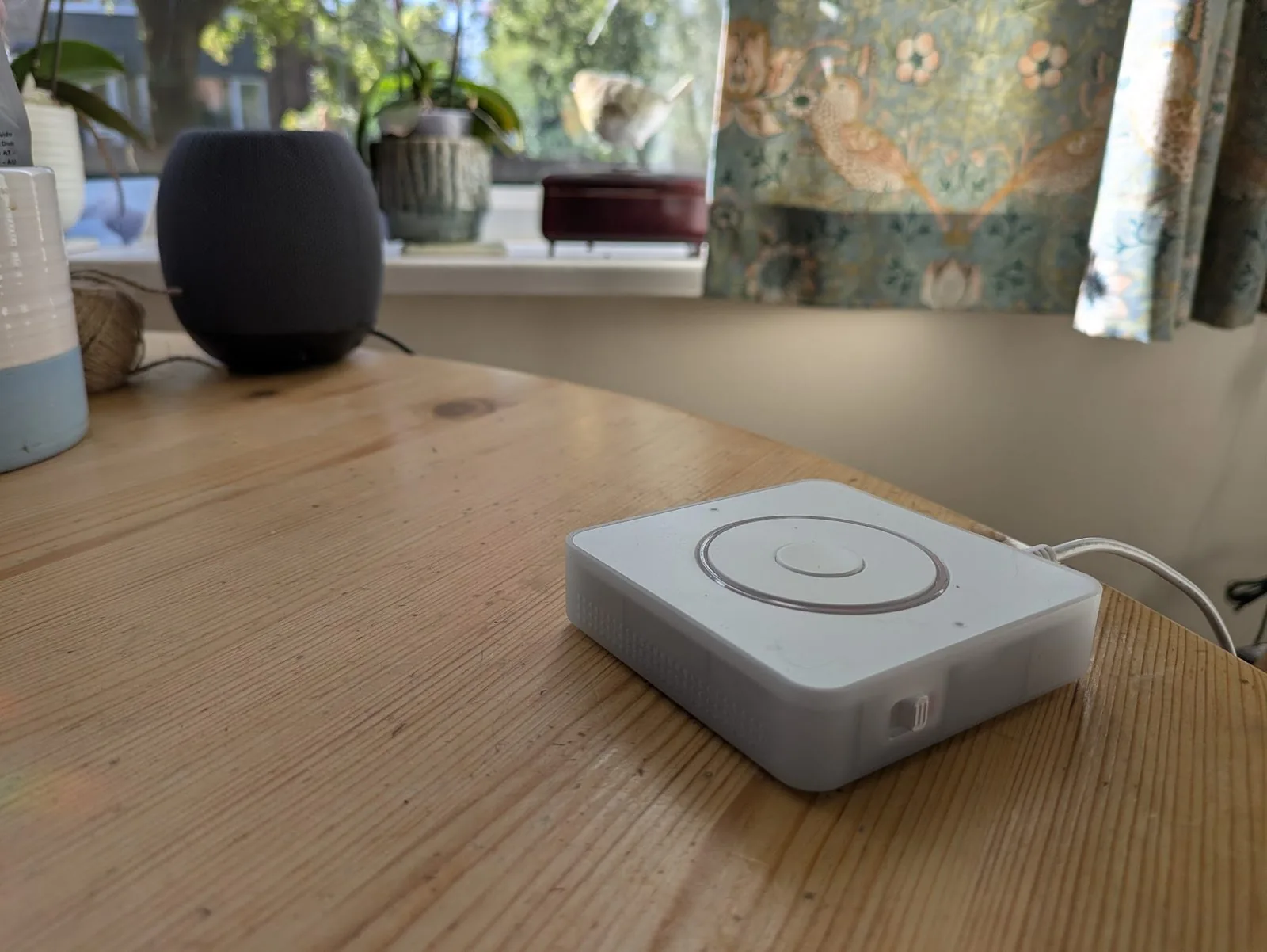 The RemindMeVoice prototype: a small white device with a circular button on top, sitting on a wooden kitchen table beside a HomePod and a sunlit window.