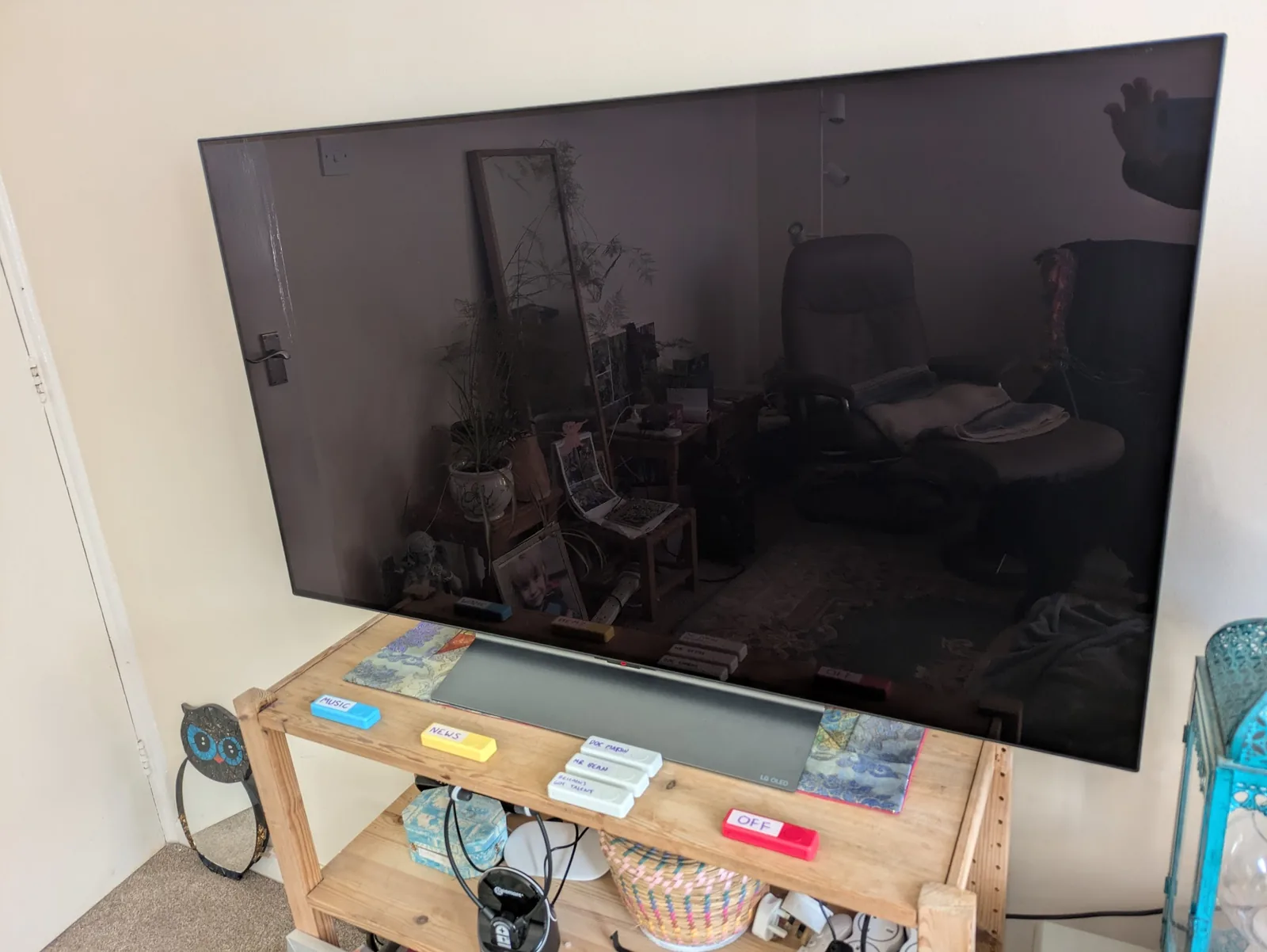 An LG TV mounted on a wall above a wooden TV stand. On the stand, six coloured Zigbee buttons are arranged in a row — blue 'MUSIC', yellow 'NEWS', three white buttons each labelled with a show name, and a red 'OFF' — alongside a soundbar and household items.